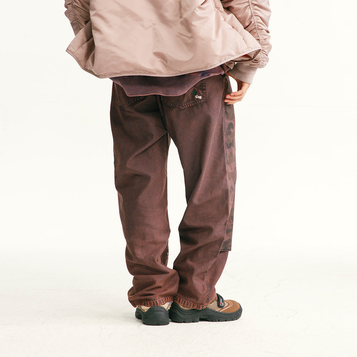 Rear view of a model wearing loose burgundy-brown pants with camouflage detail along the outer leg.