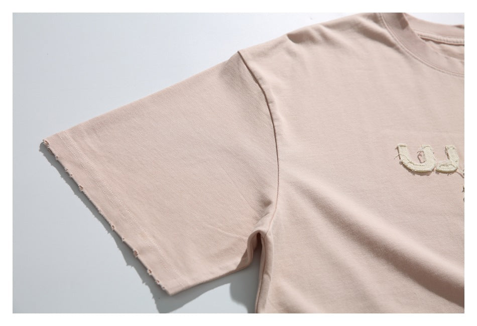 Close-up of a pale blush-pink T-shirt sleeve with a raw, frayed edge and part of the chest lettering.