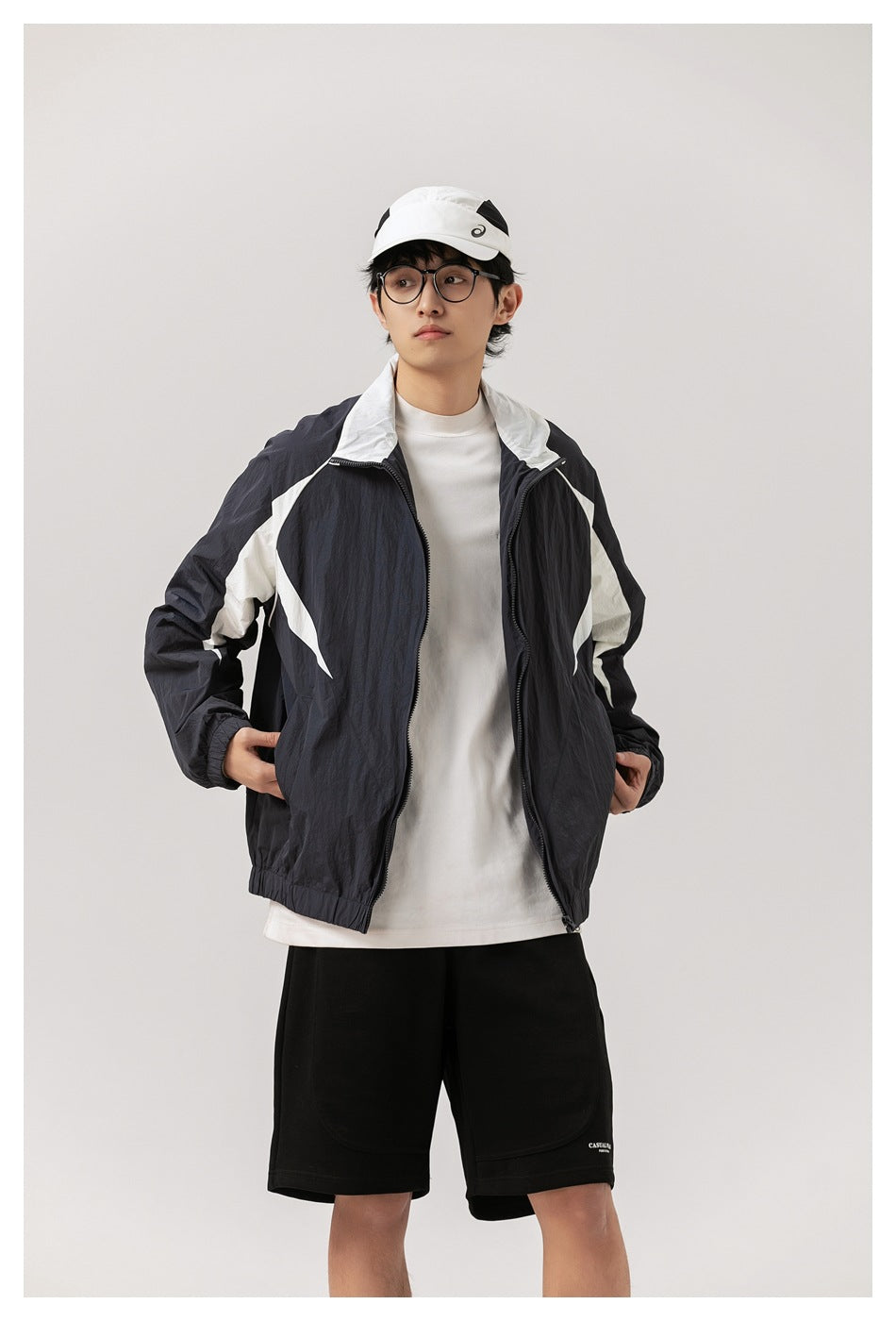 Model wearing the navy and white color-block jacket open over a white tee with black shorts and a white cap