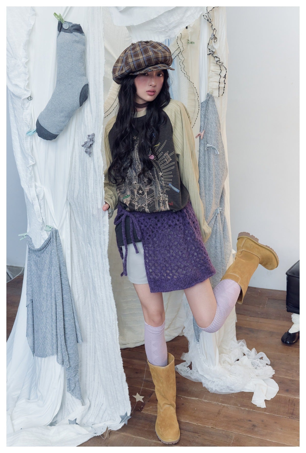 Model wearing a charcoal graphic raglan top with cream ribbed sleeves, a purple wrap skirt, plaid cap and boots.