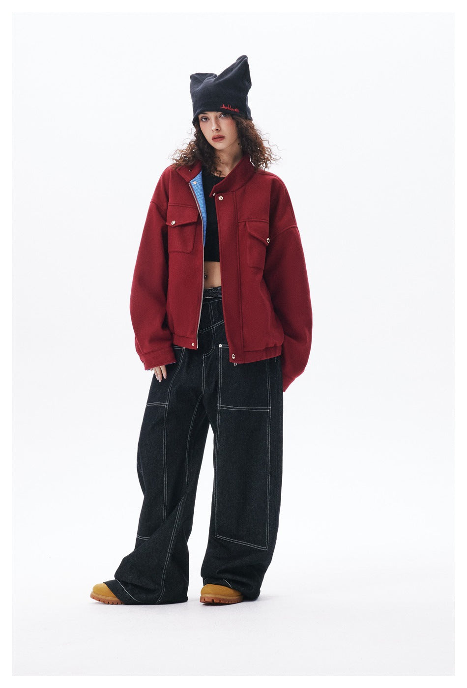 Model wearing the red jacket open over a black top with wide dark jeans, a dark beanie and yellow boots