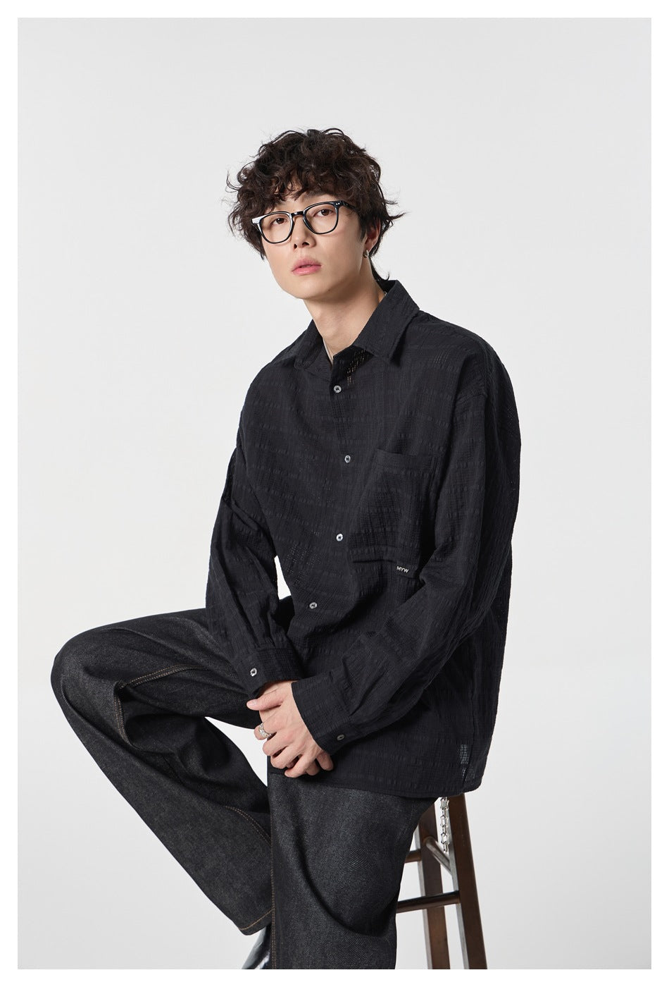 Model seated on a stool wearing a black textured check button-up shirt with dark wide-leg jeans, three-quarter view