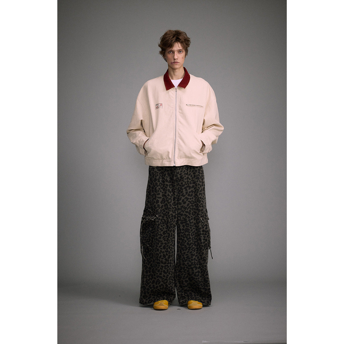 Model wearing a cream zip-front jacket with a dark red collar and chest pockets, paired with patterned wide-leg trousers.