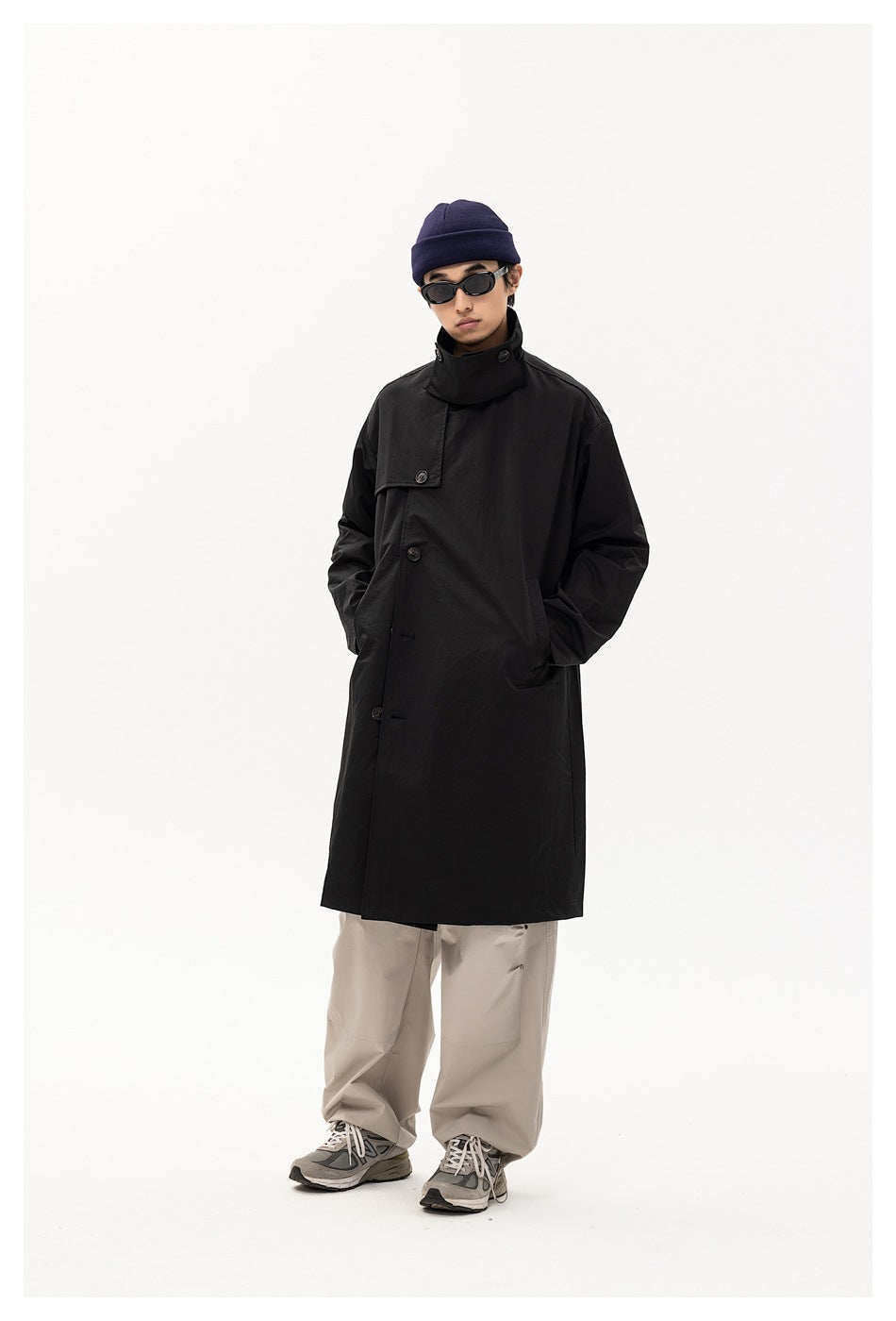 Full-length front view of a model wearing a long black high-collar coat with loose light-gray trousers.