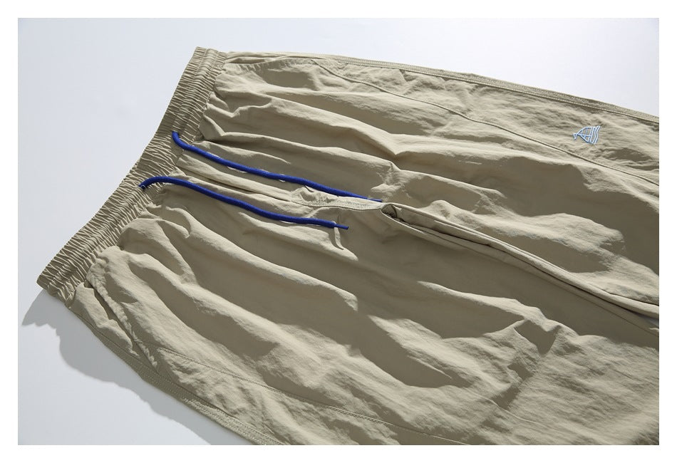 Angled close-up of beige jogger pants with an elastic waistband, blue drawstrings and a small embroidered emblem.