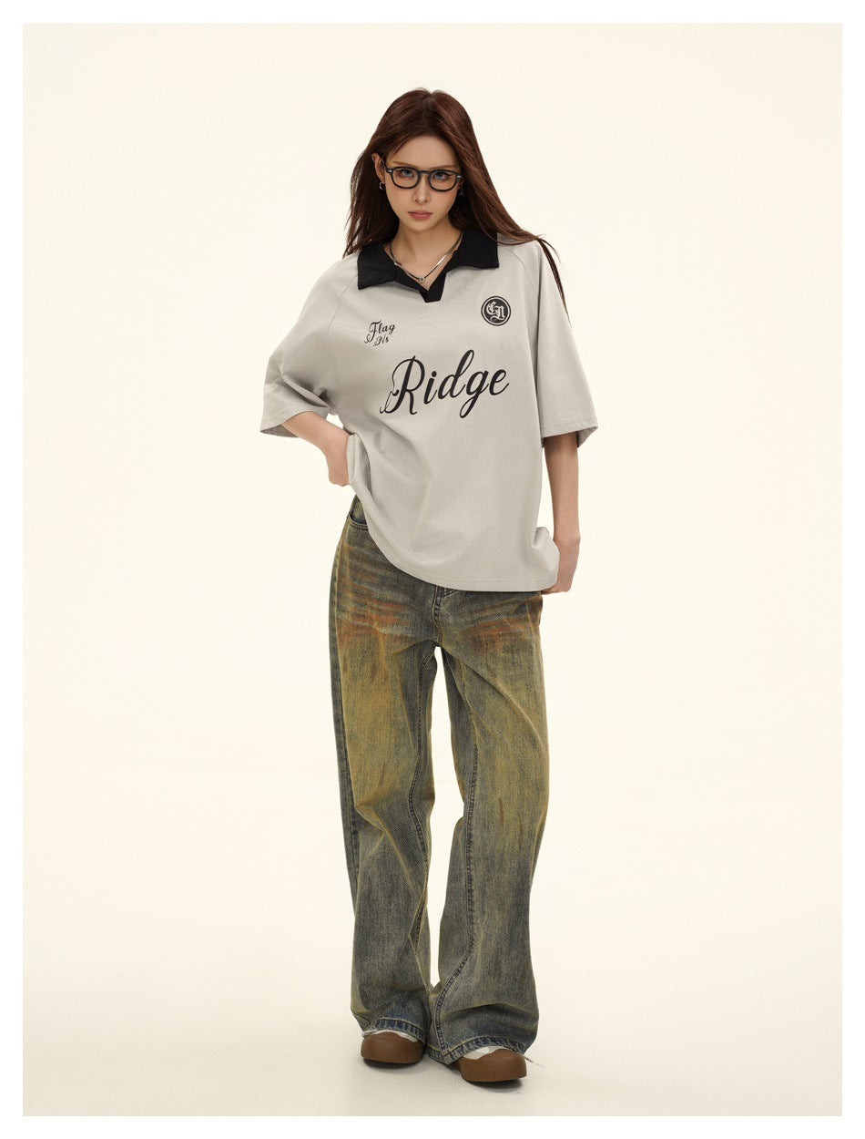 Full-length model wearing a light gray oversized polo with black collar and lettering, and loose faded olive-gray jeans.