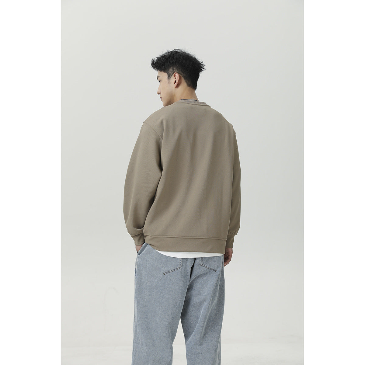 Back view of a man wearing a plain taupe crewneck sweatshirt and light blue jeans.