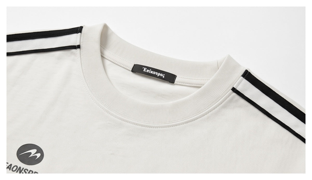 Close-up of a white crew neck, Greek-letter neck label, black shoulder stripes, and partial chest graphic.