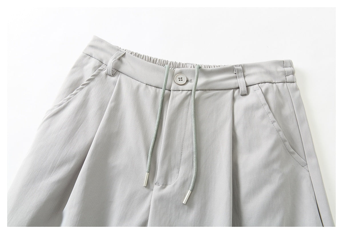 Close-up of light gray trousers showing a buttoned waistband, pleats, belt loops, slanted pockets and long drawstrings.