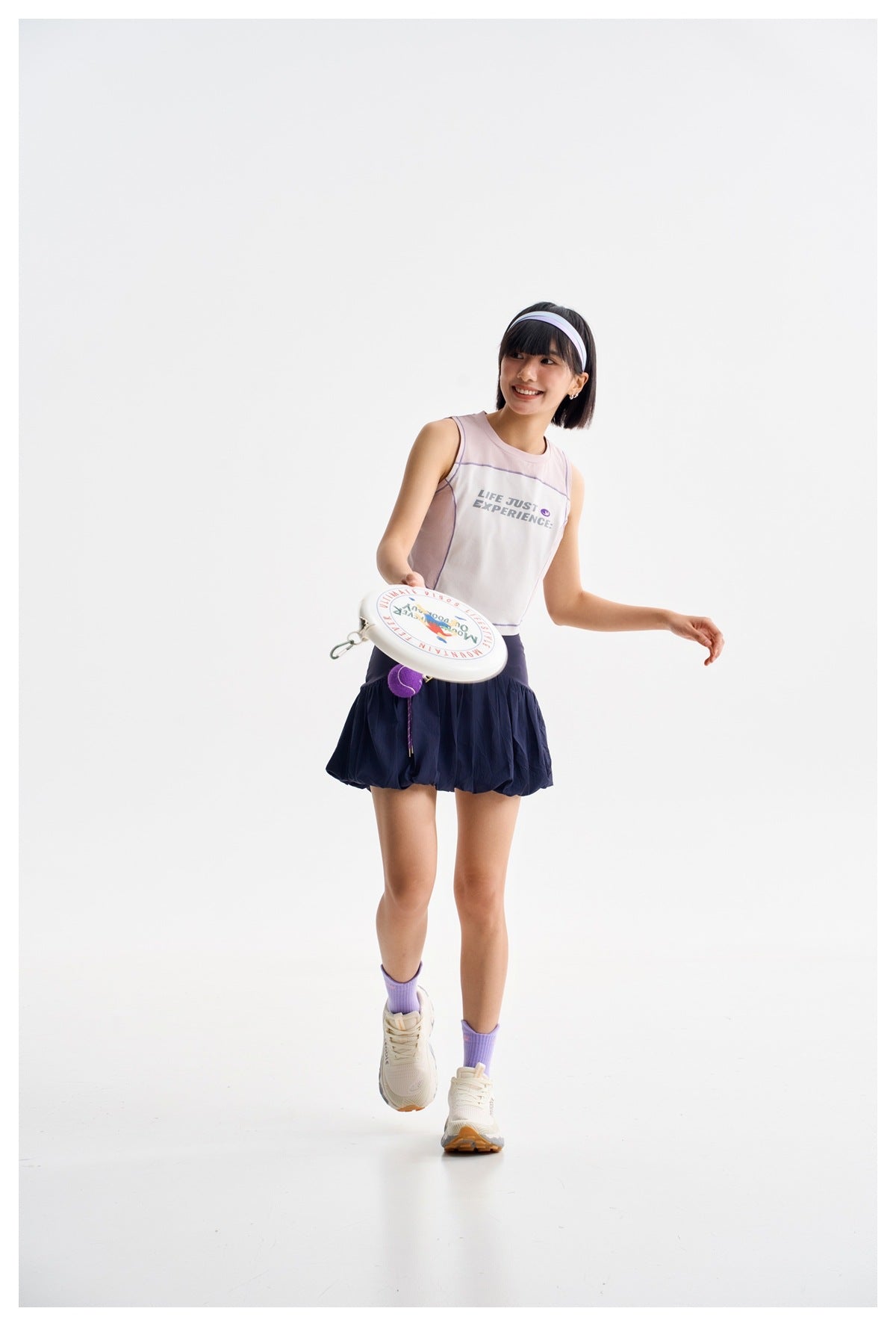 A smiling model wears a pale pink sleeveless crop top and navy bubble skirt while holding a flying disc.