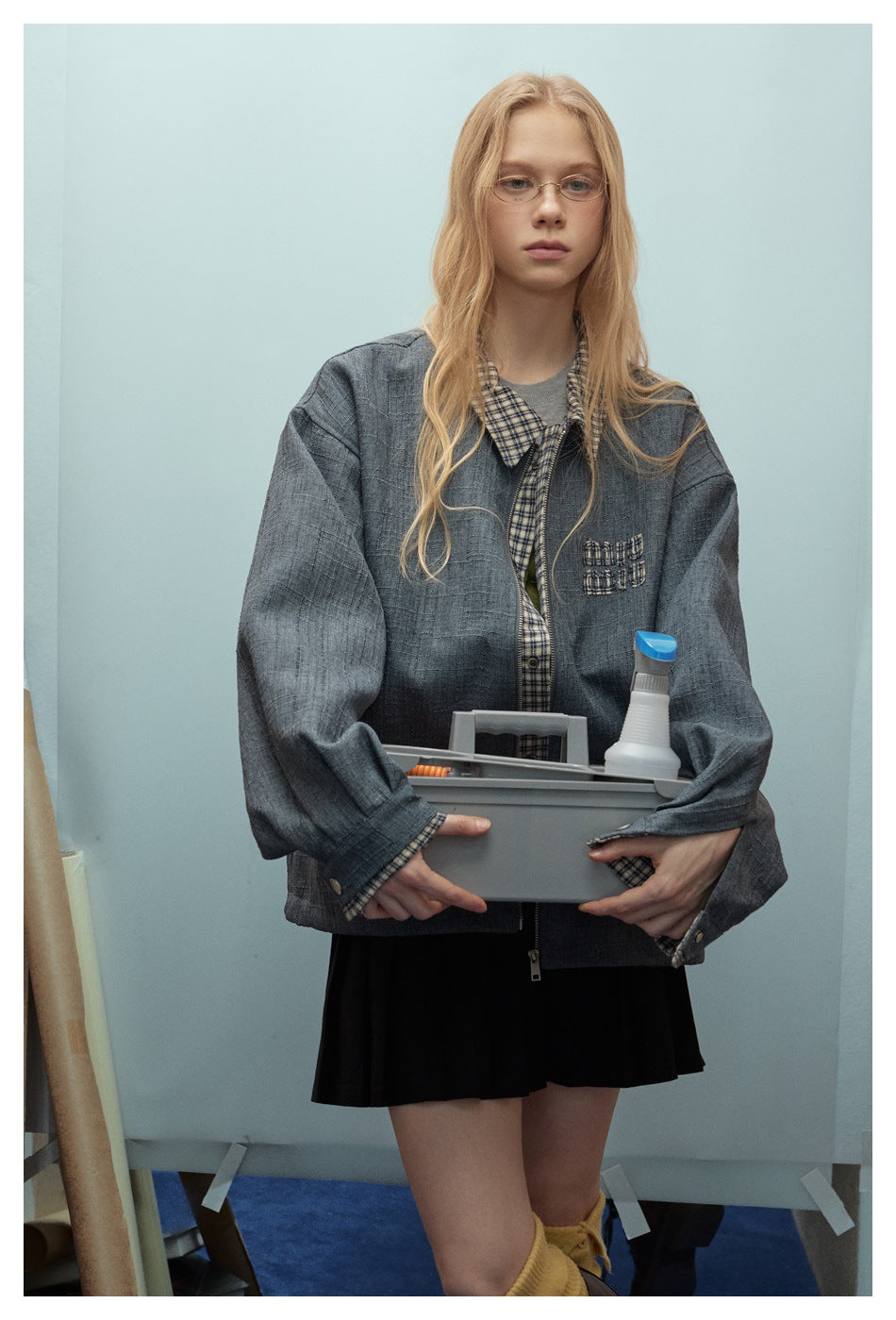 Front view of a model wearing an oversized charcoal jacket with checked trim and chest embroidery, holding a gray caddy.