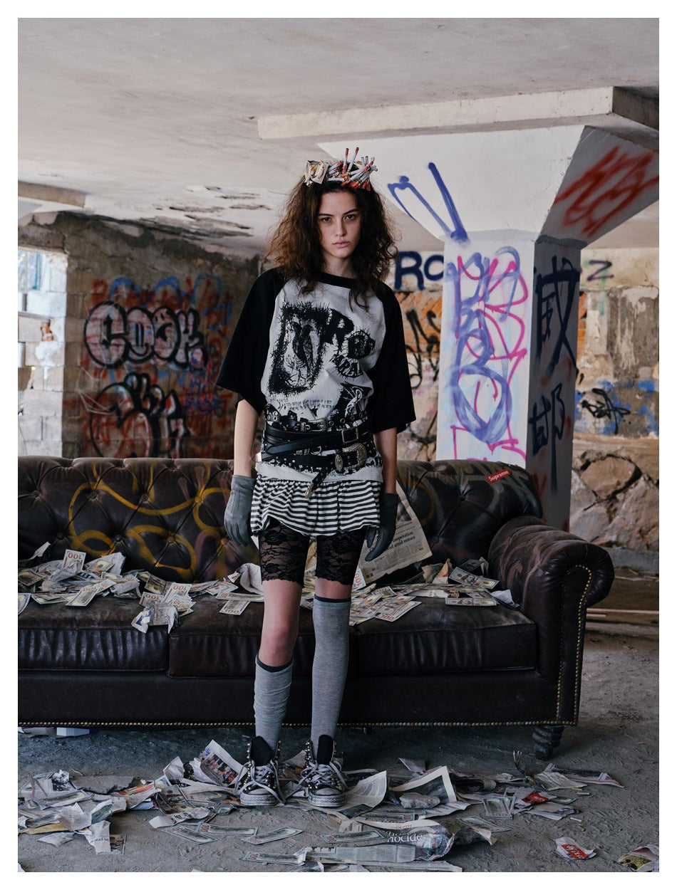 Model wears a black-sleeved graphic raglan shirt with a striped skirt and layered belts, standing before a dark sofa.