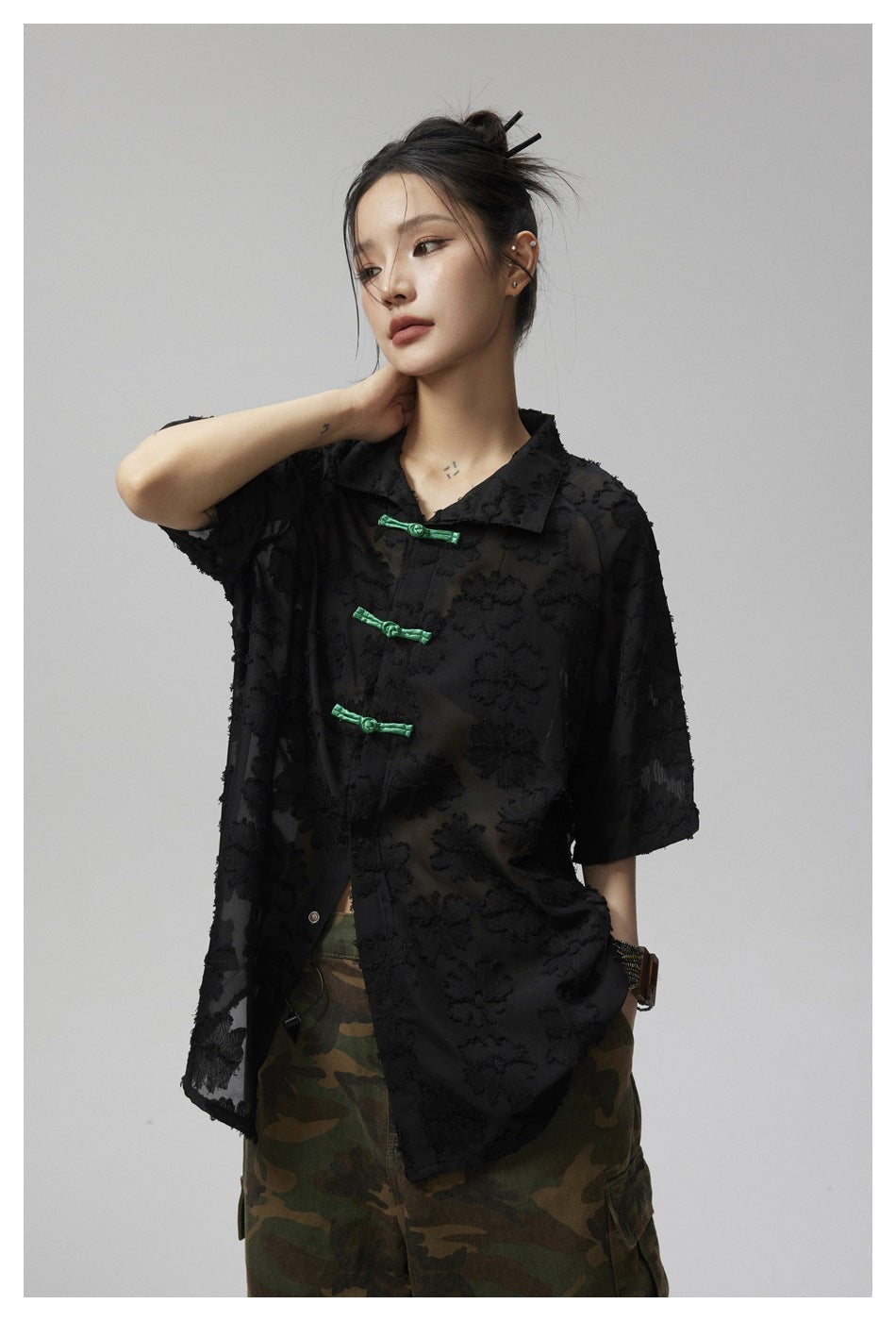 Model wearing a black sheer floral-textured short-sleeve shirt with green frog closures and camouflage trousers.