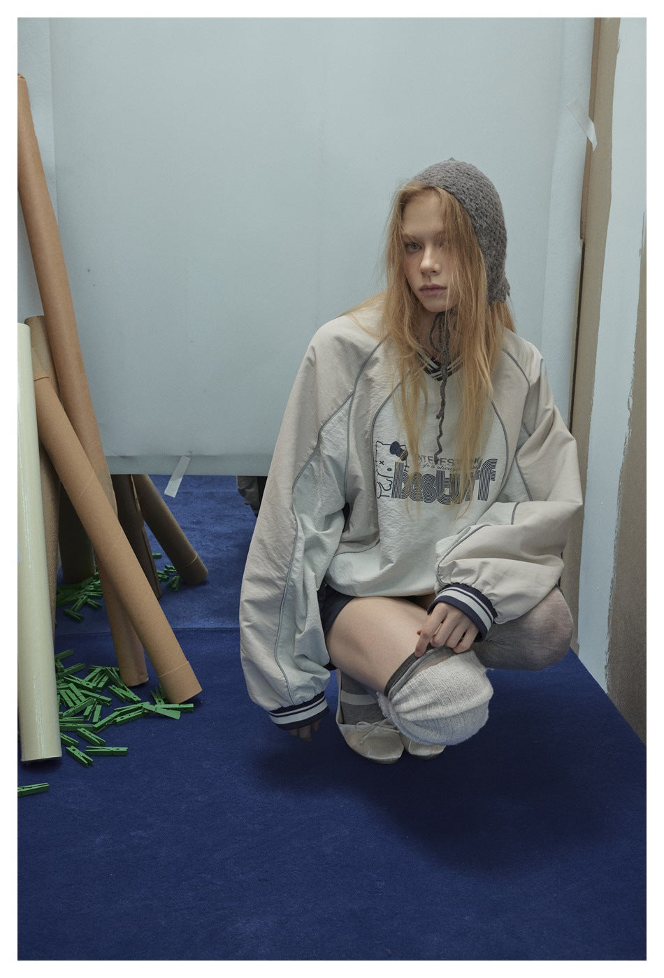 Seated model wearing a cream graphic pullover with gray panels and striped trim, paired with shorts and leg warmers.