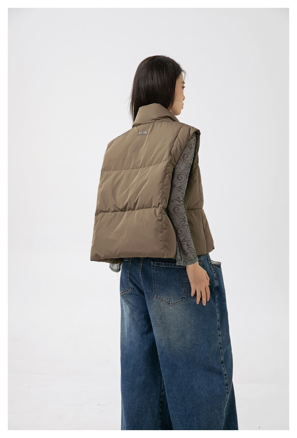 Model wearing the back of a taupe quilted sleeveless puffer vest with a high collar and wide padded panels.