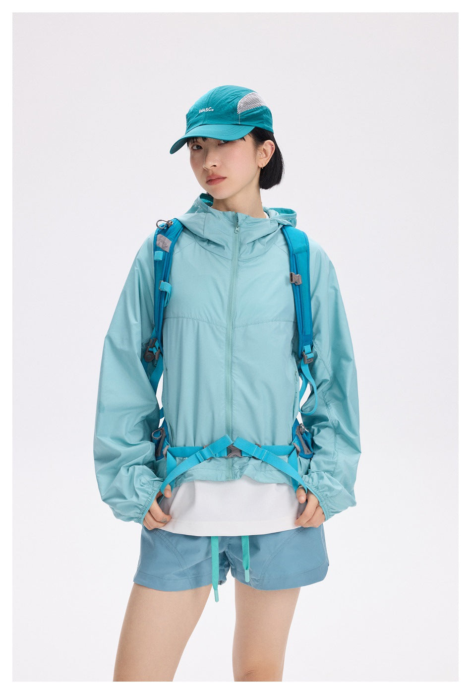 Model wearing a pale aqua hooded jacket, blue hiking backpack, teal cap, and matching shorts, front view.
