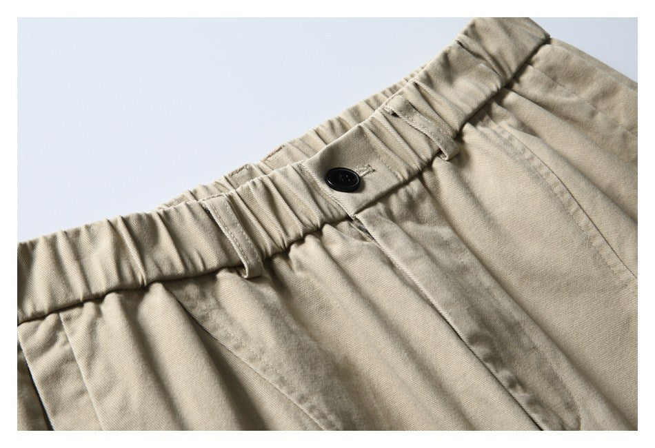 Close-up of light beige trousers showing the gathered waistband, belt loops, black button, and front pocket seam.