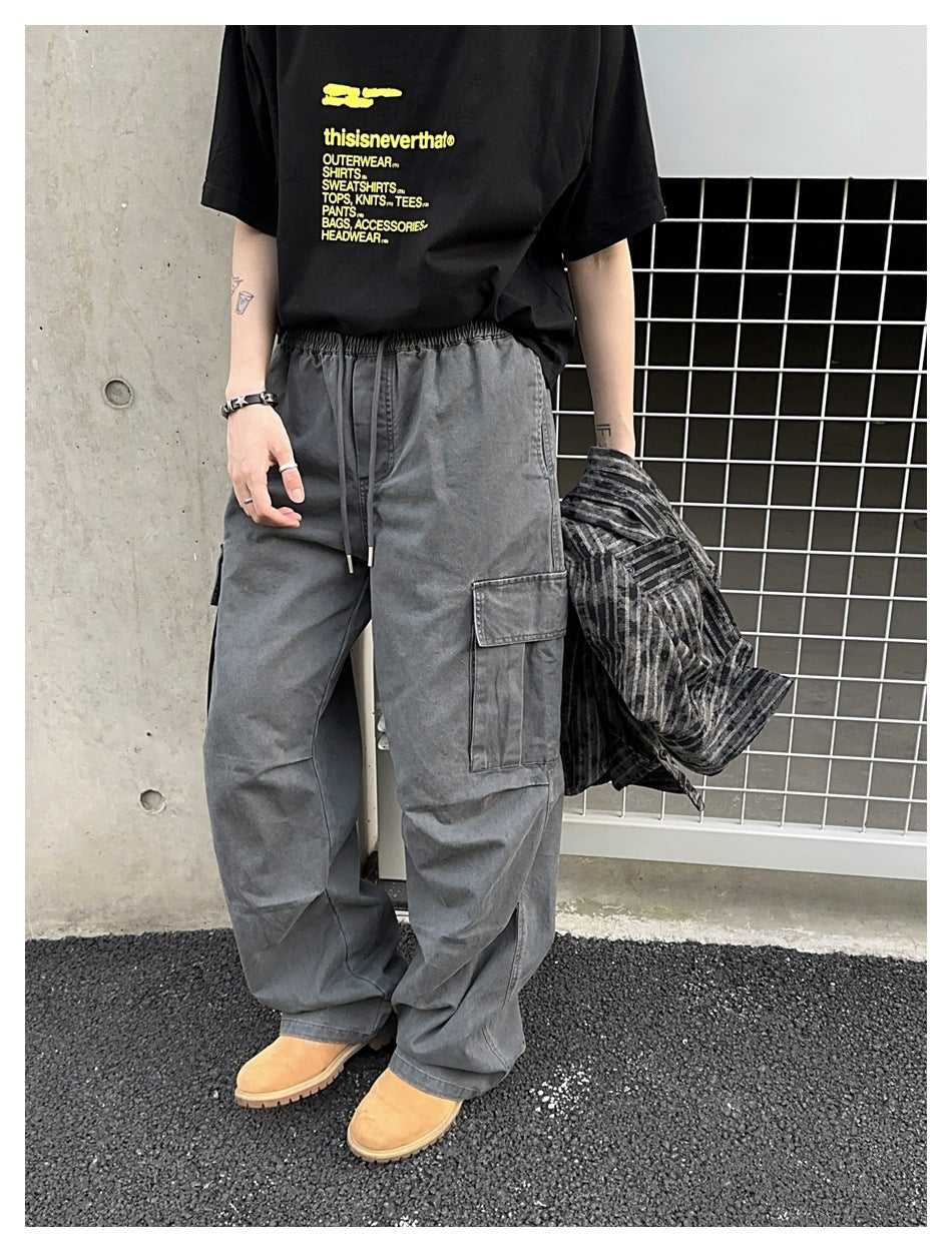 Model wearing loose gray cargo pants with a drawstring waist and thigh pocket, holding a dark striped shirt.