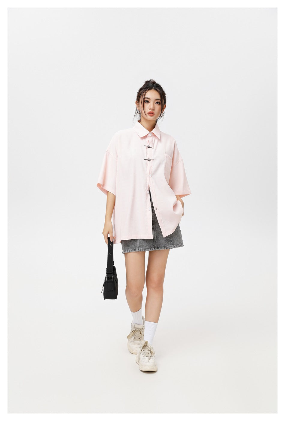 Model wearing a pale pink oversized short-sleeve shirt with decorative front clasps and a chest pocket, paired with a gray