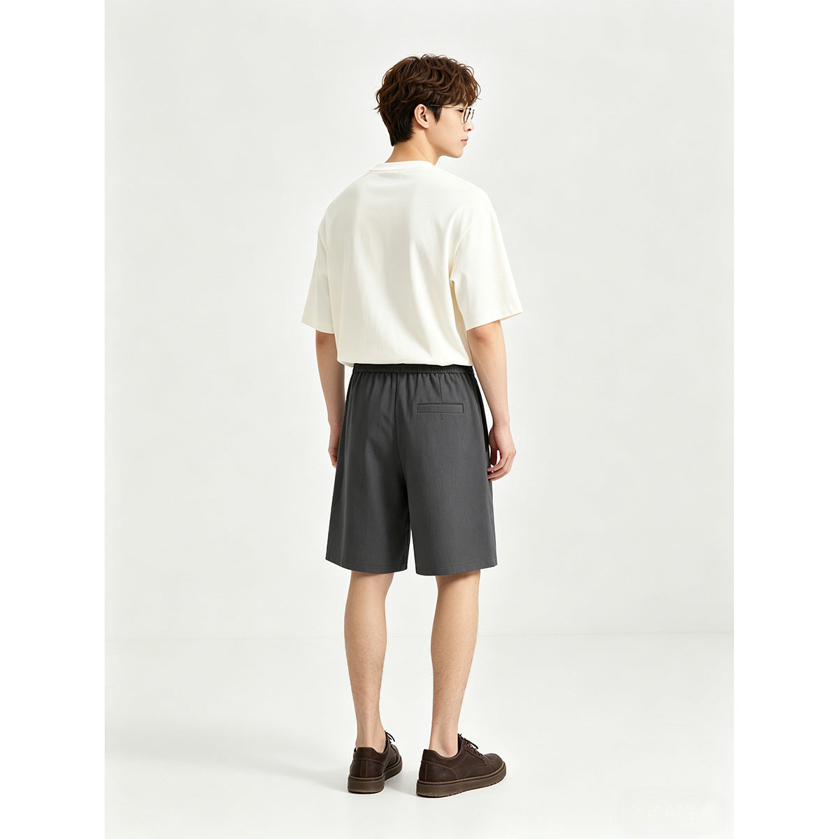 Rear view of a model wearing a white T-shirt and charcoal shorts with an elastic waistband and back pocket.