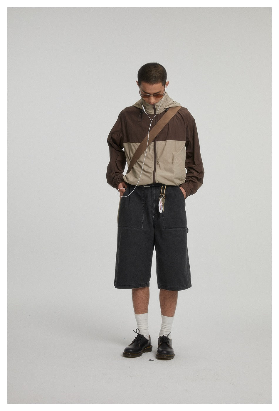 Full-length view of model in brown and khaki hooded zip jacket with crossbody strap, dark shorts and black shoes