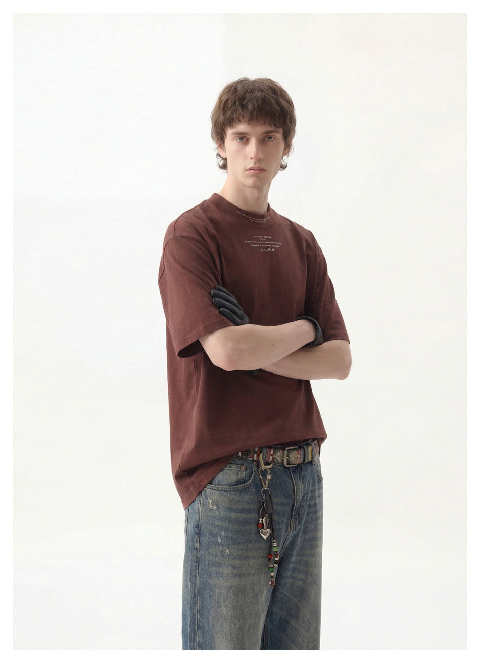 Model wearing a dark brown short-sleeve T-shirt with small chest lettering and blue jeans.