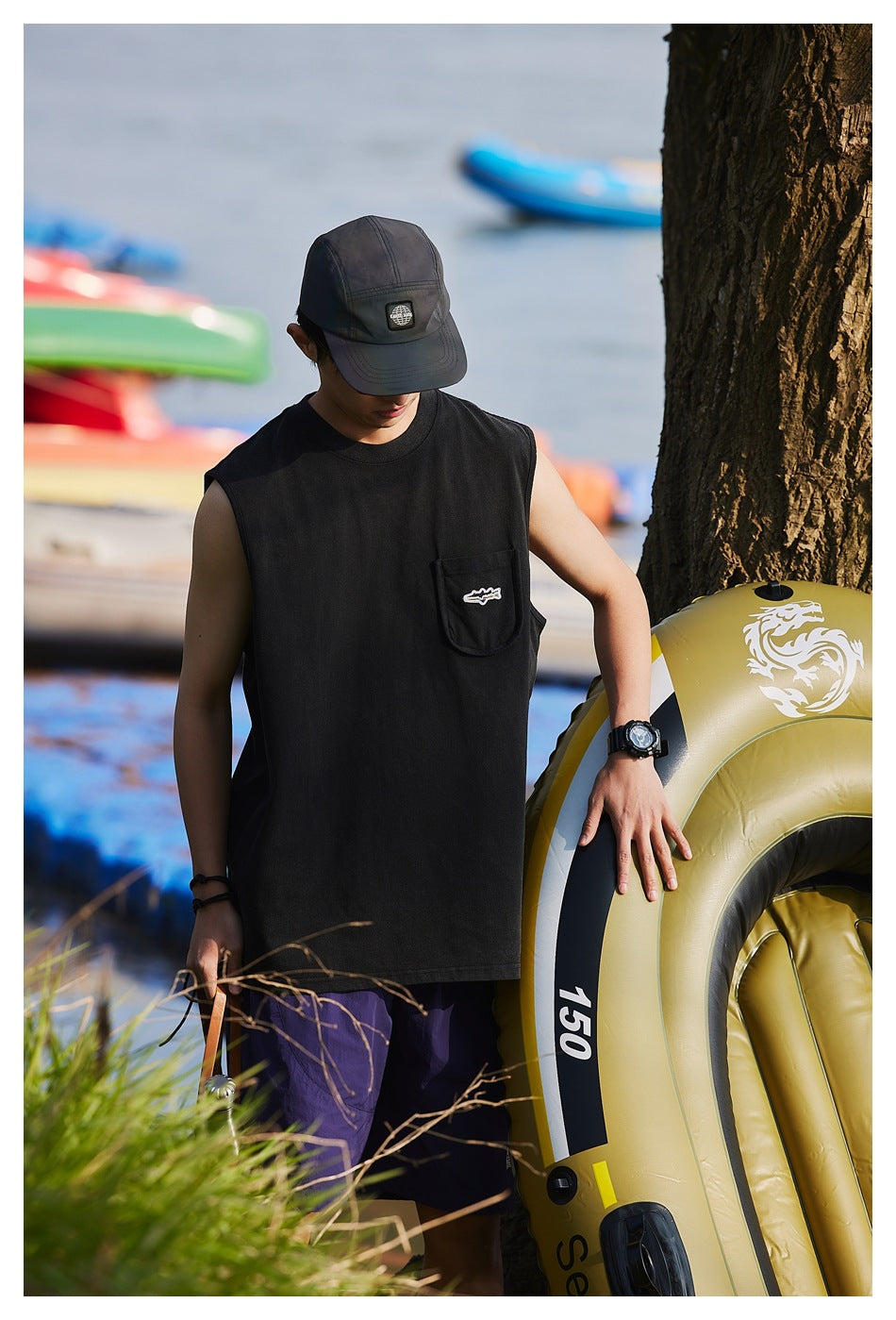 Model wearing a black sleeveless pocket tank top with a cap and purple shorts beside an inflatable boat