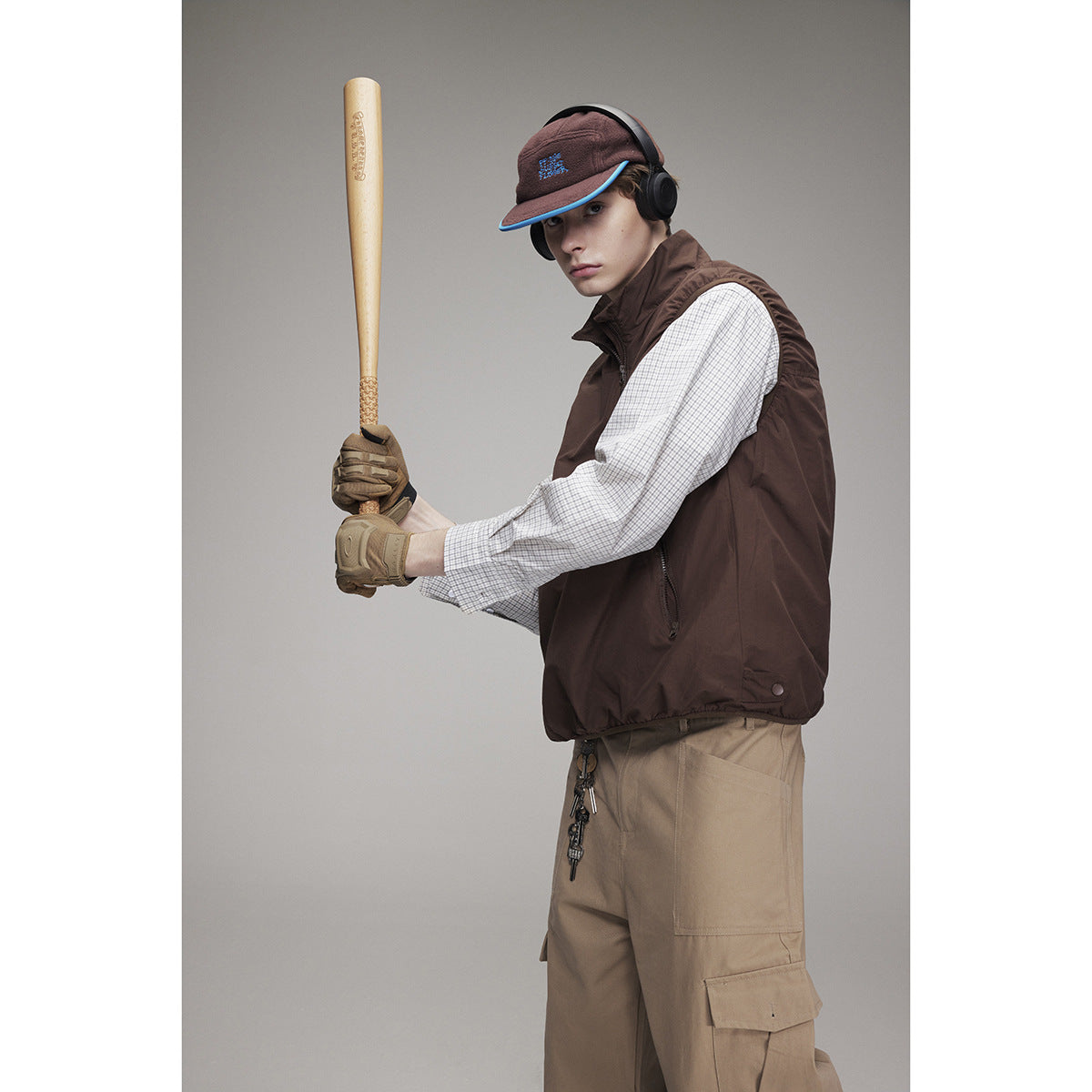 Model wearing a dark brown sleeveless zip vest over a checked shirt, with beige cargo pants and a cap.