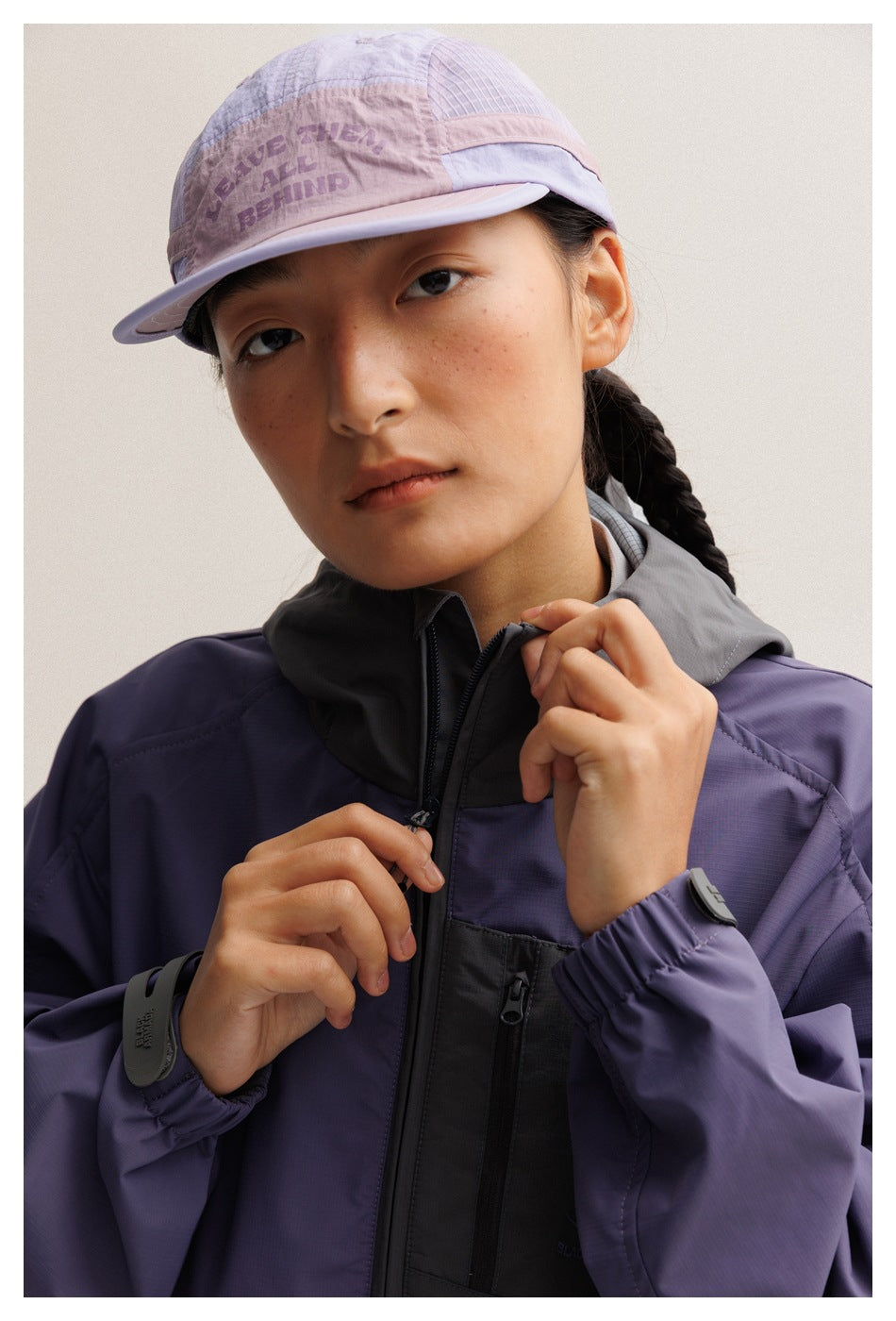 Close-up of a model wearing a purple jacket with a charcoal hood and zip pocket, and a lavender cap with printed lettering.