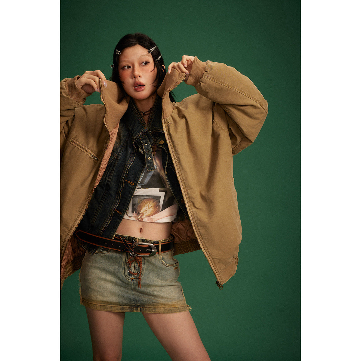 Model wearing an oversized tan zip-front jacket open over a dark denim vest and graphic top.