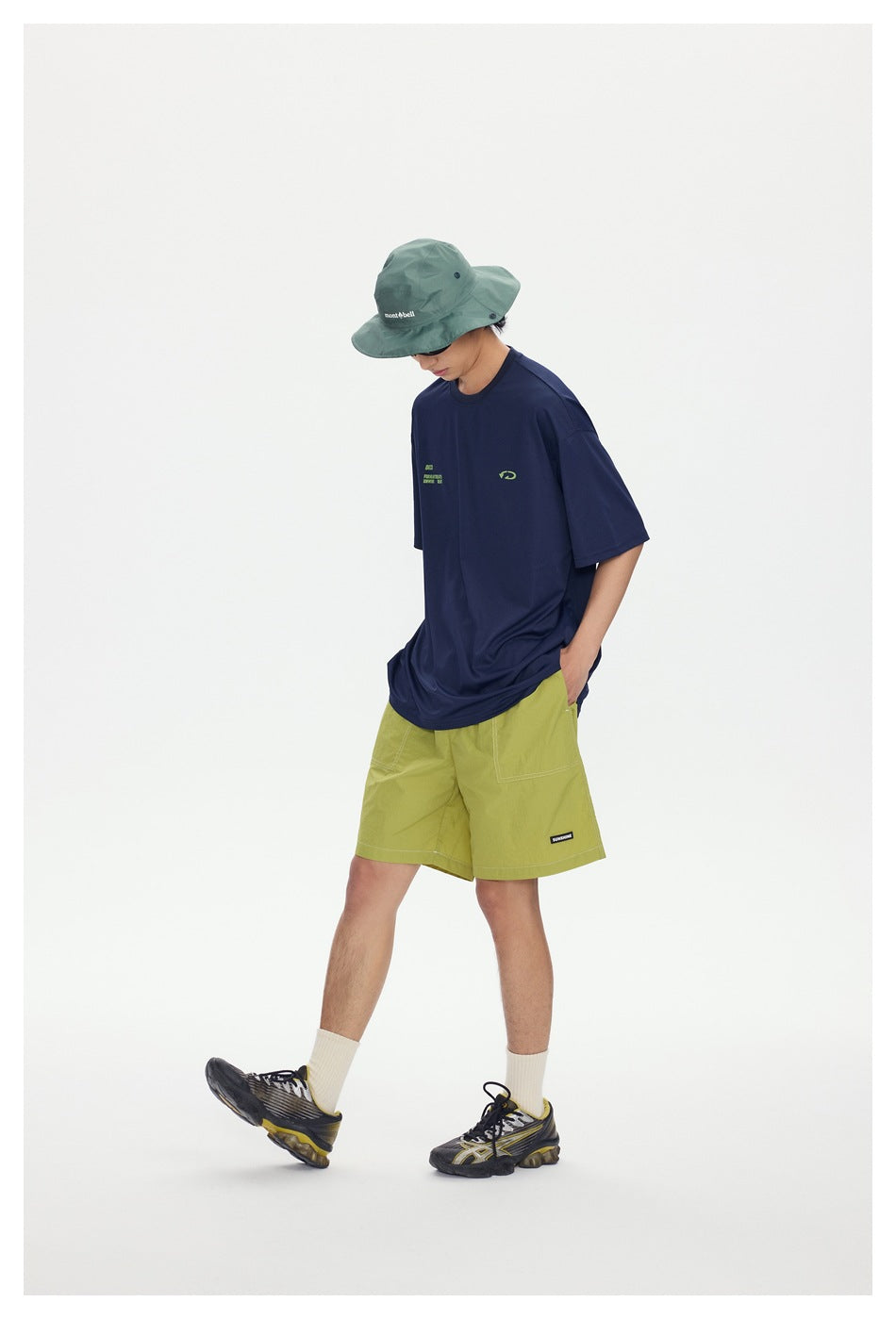 Model in a navy graphic T-shirt and lime shorts, wearing a green bucket hat and stepping forward in dark sneakers.
