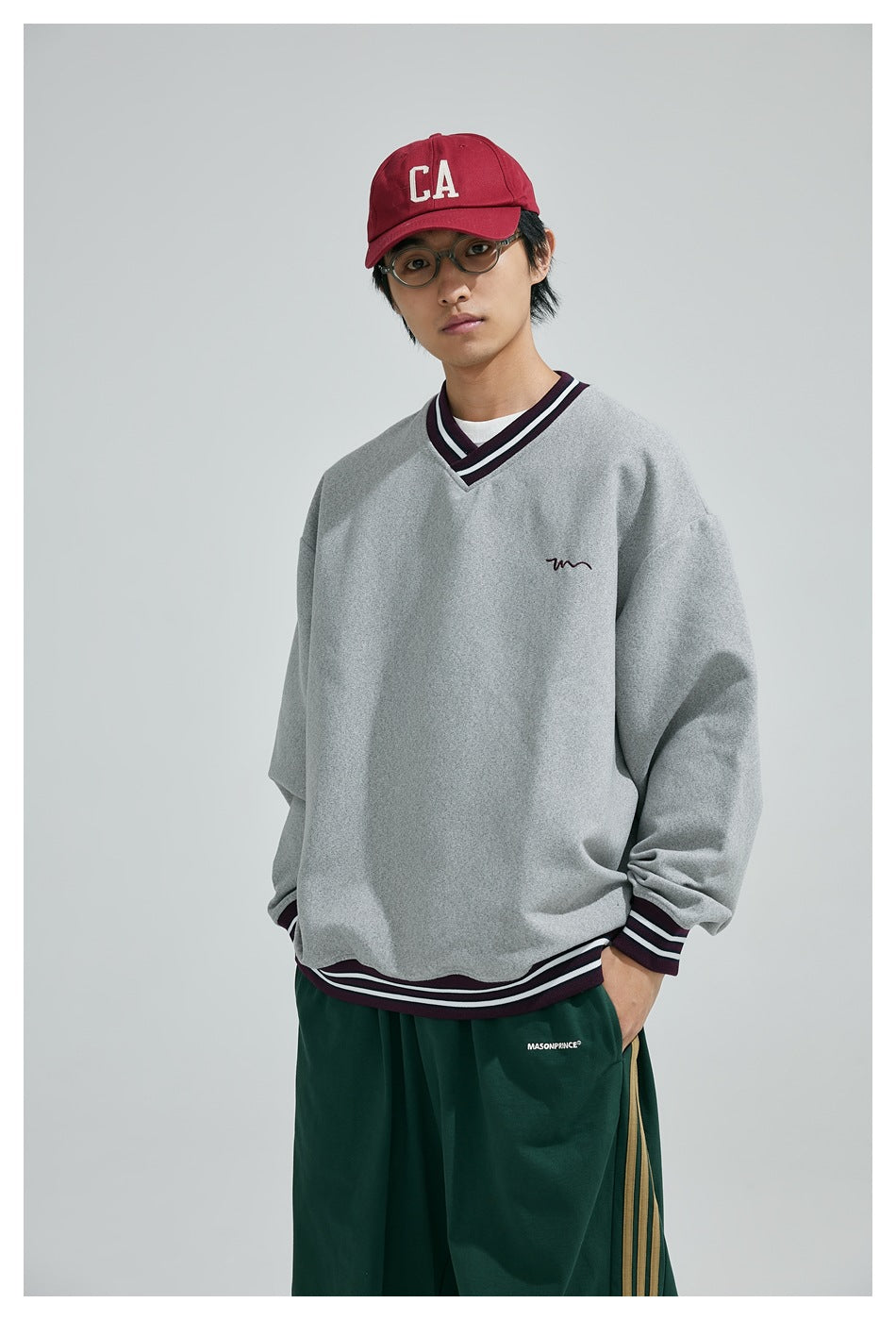 Model wearing a gray oversized V-neck sweatshirt with burgundy striped trim and dark green pants.