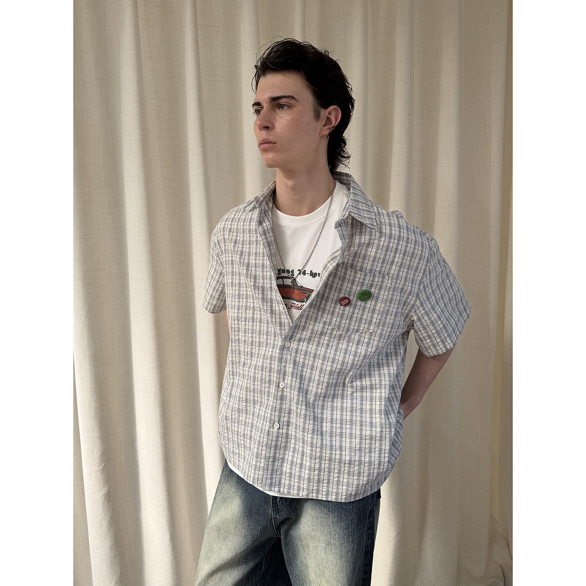 Closer front view of a man wearing an open blue-gray plaid short-sleeve button-up shirt over a white graphic tee.