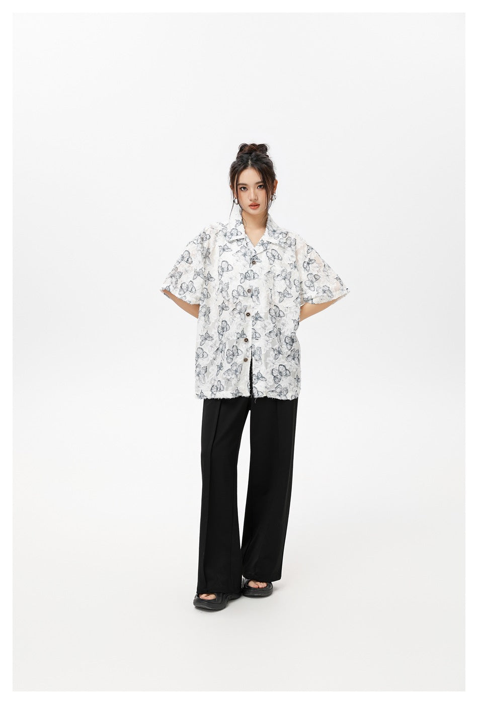 Model wearing a loose white short-sleeve shirt with gray butterfly print and black wide-leg pants, posed with arms behind