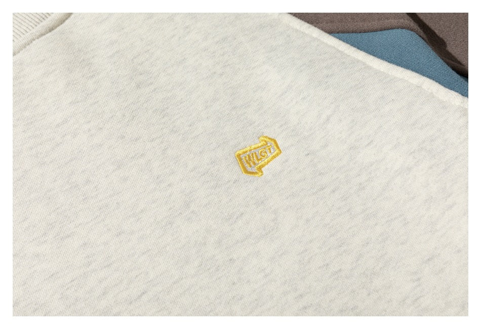 Close-up of a small gold embroidered emblem on an off-white heathered sweatshirt.