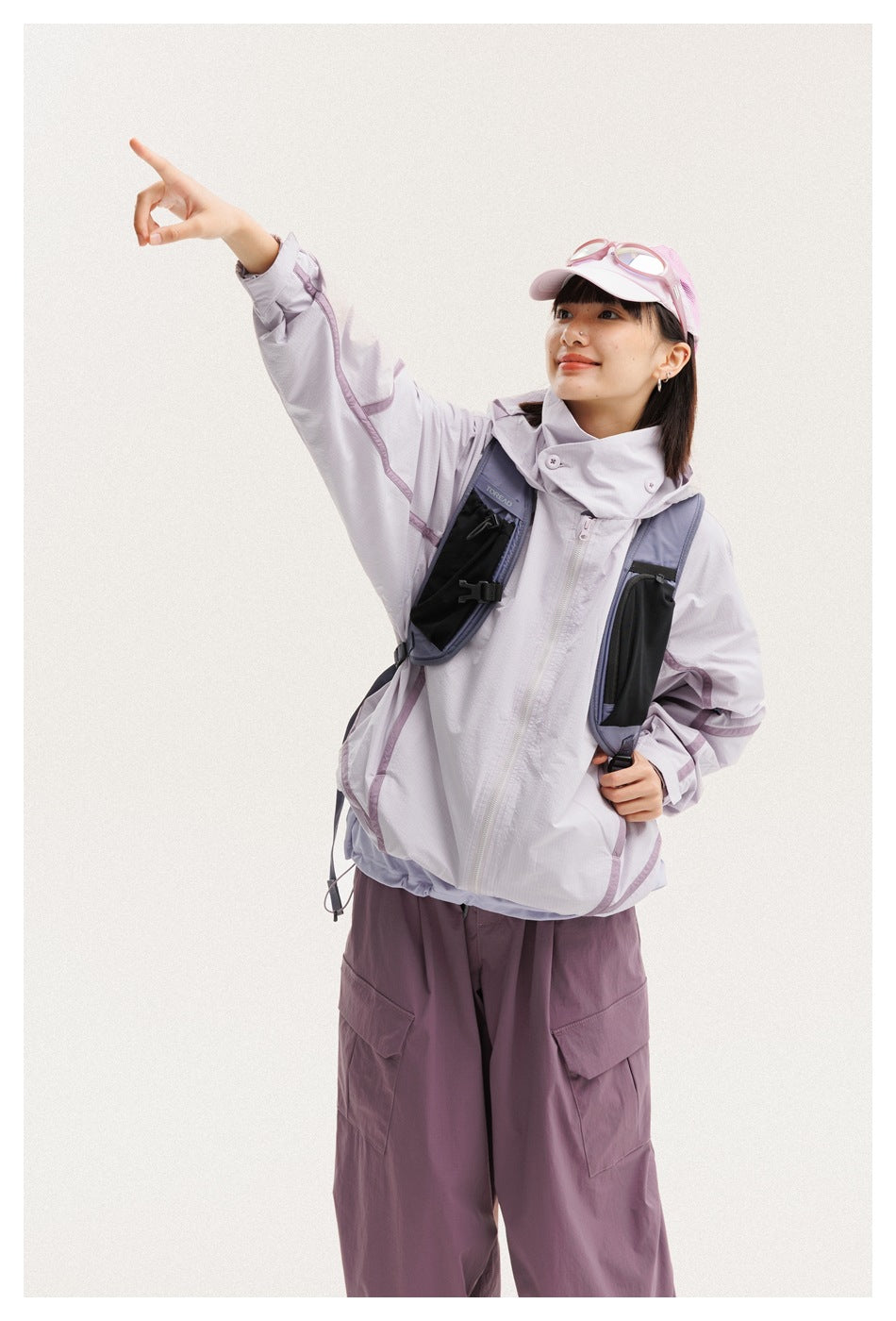 Woman in a light lavender zip jacket and plum cargo pants, wearing a pink cap and a black backpack.
