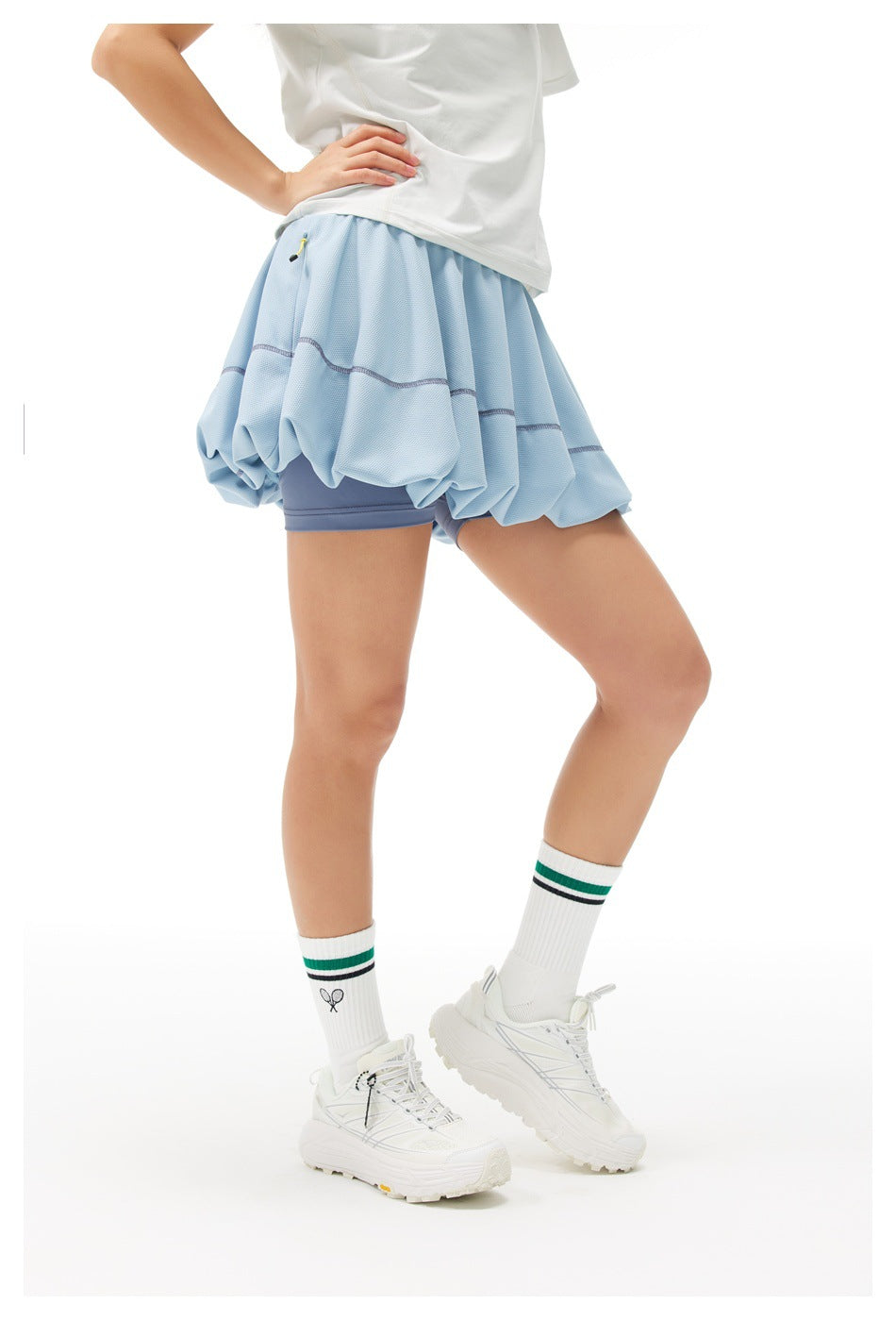 Model in a light blue bubble tennis skirt with dark blue built-in shorts, shown at an angled front view.