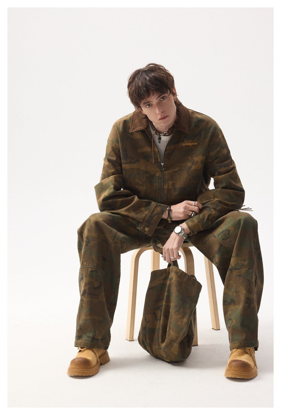 Seated model wearing a brown-and-olive camouflage zip jacket with matching loose trousers and a dark brown collar.