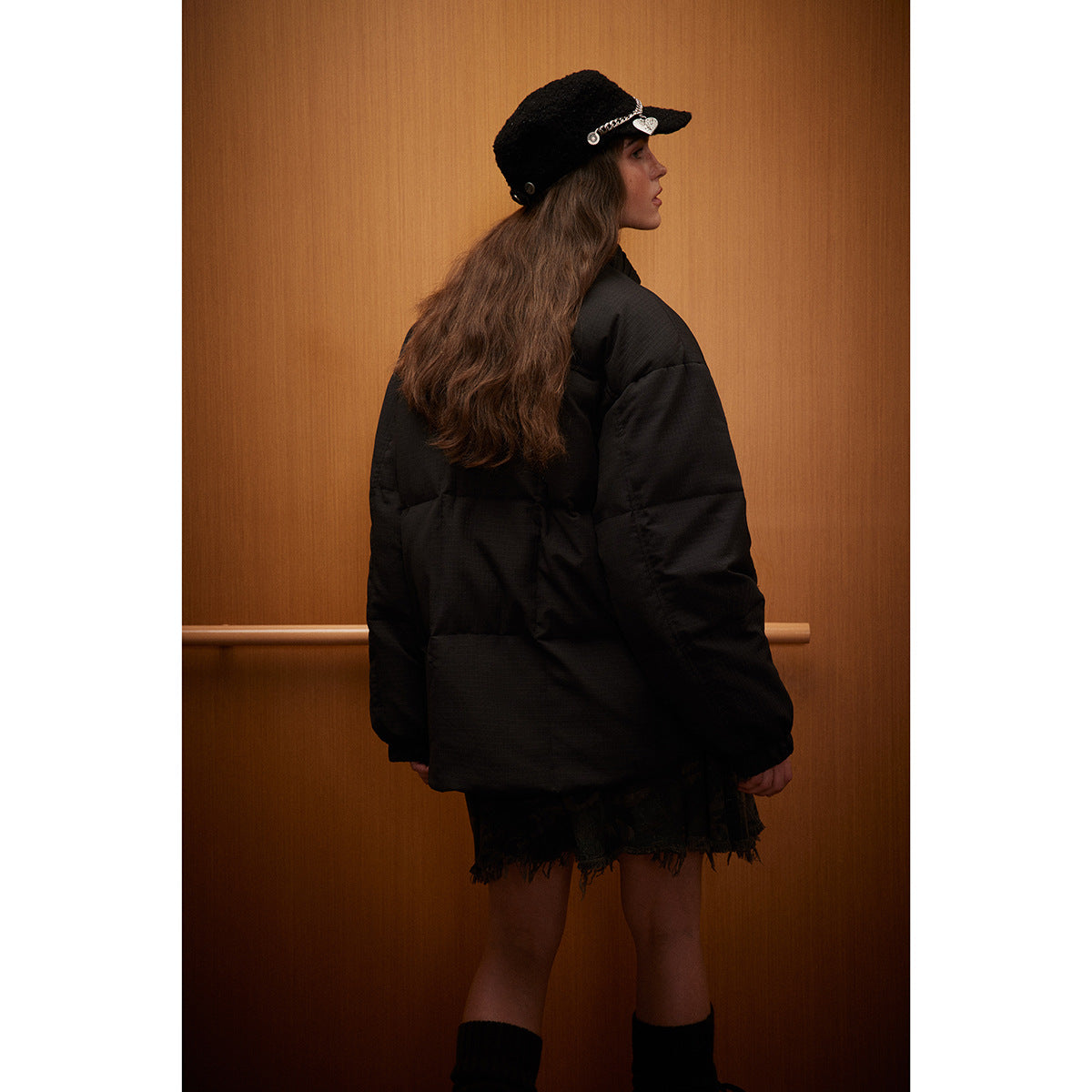 Rear view of a model wearing a black padded jacket, with a black cap and a dark frayed skirt.