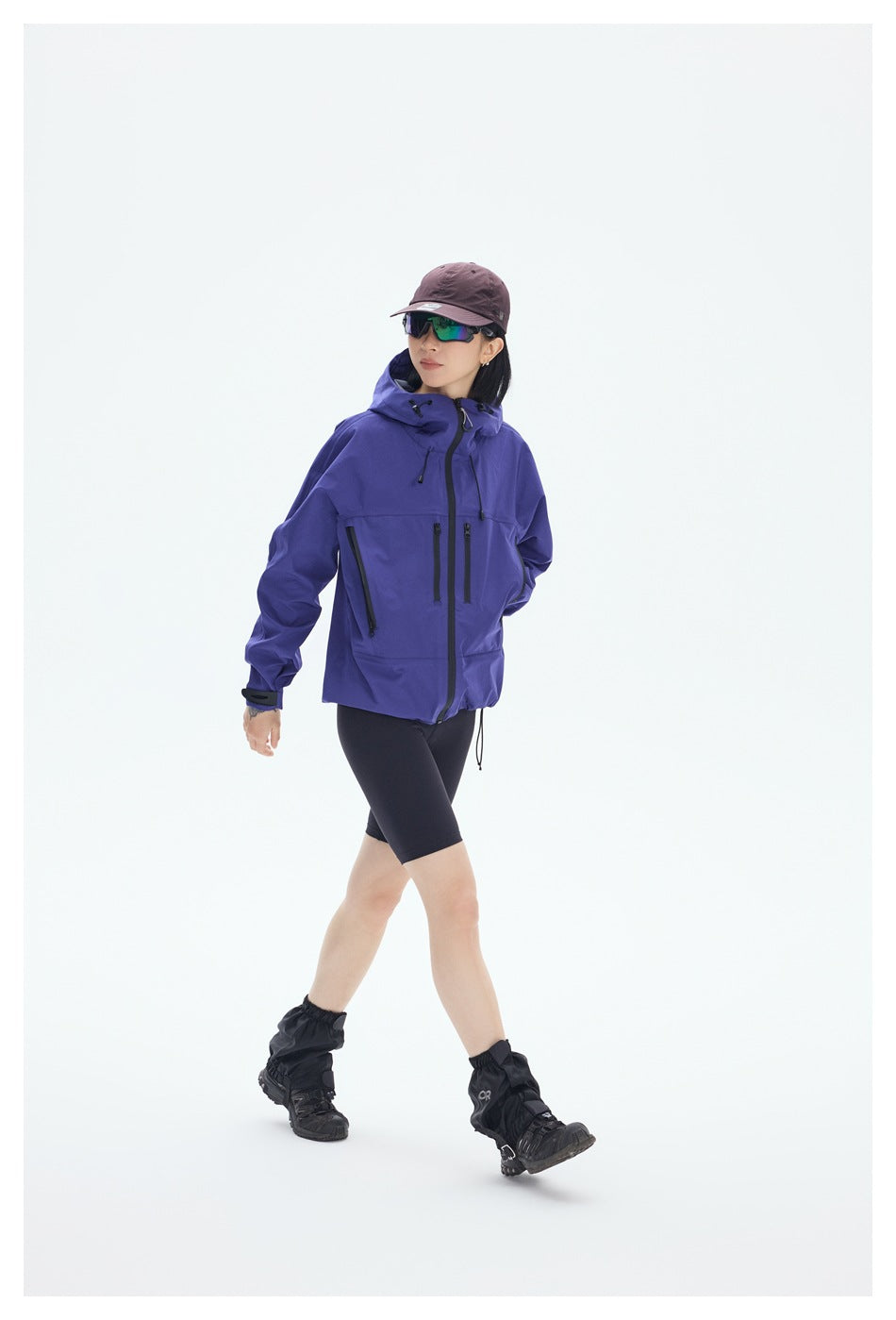 Model walking in a purple hooded jacket with black zip pockets, black shorts, and hiking gaiters.