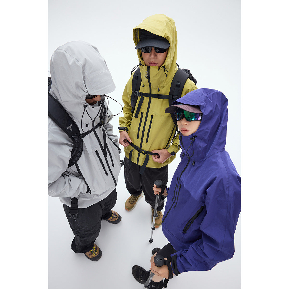 Three hikers wear gray, yellow, and violet hooded jackets with backpacks and trekking poles.