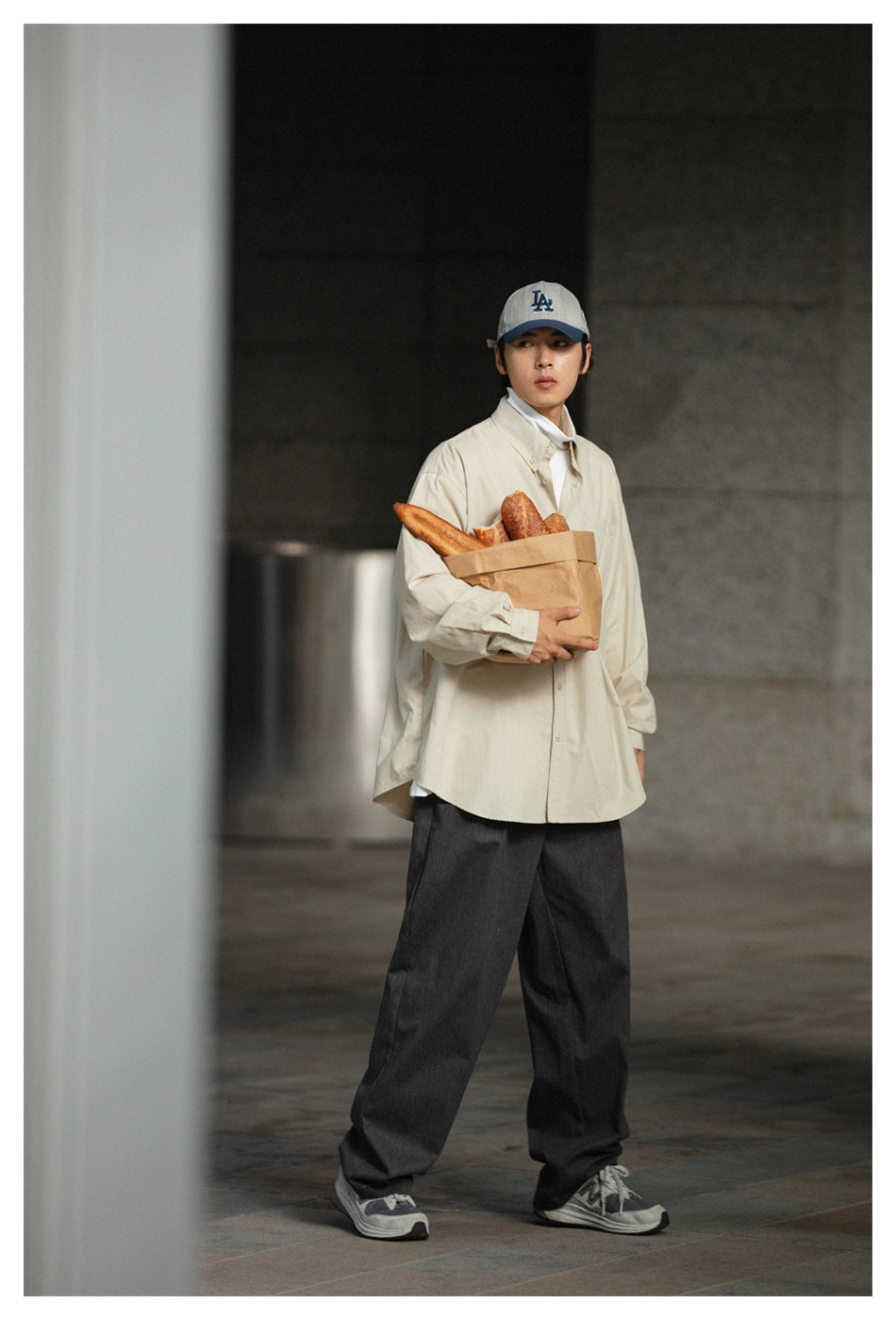 Man wearing a cream oversized button-down shirt over a white top with dark trousers, holding a bag of bread