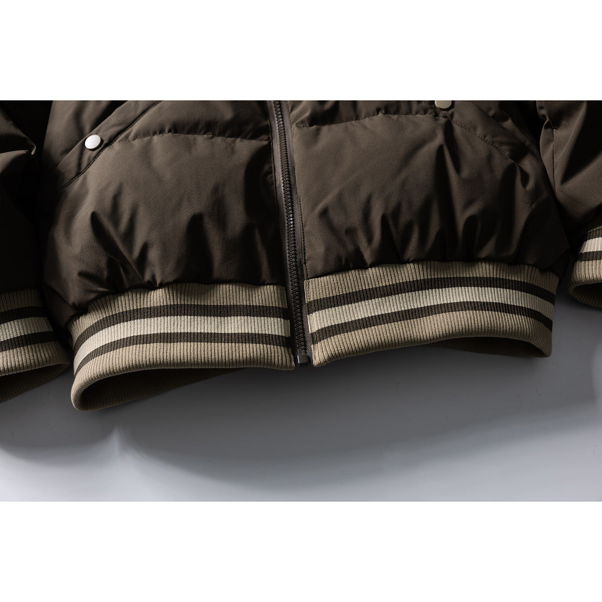 Close-up of a dark brown padded jacket's front zipper, lower hem, and striped rib-knit waistband.