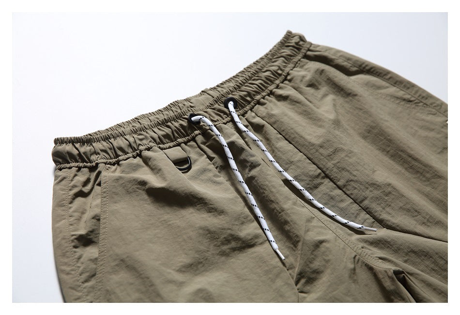 Close-up of taupe shorts showing the elastic waistband, white drawstrings, black D-ring, and front pocket seams.