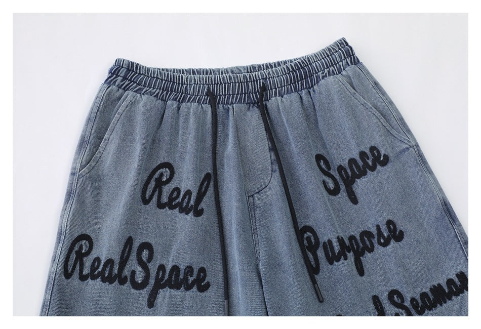 Cropped front view of blue denim shorts with an elastic waistband, black drawstrings, pockets, and script lettering.