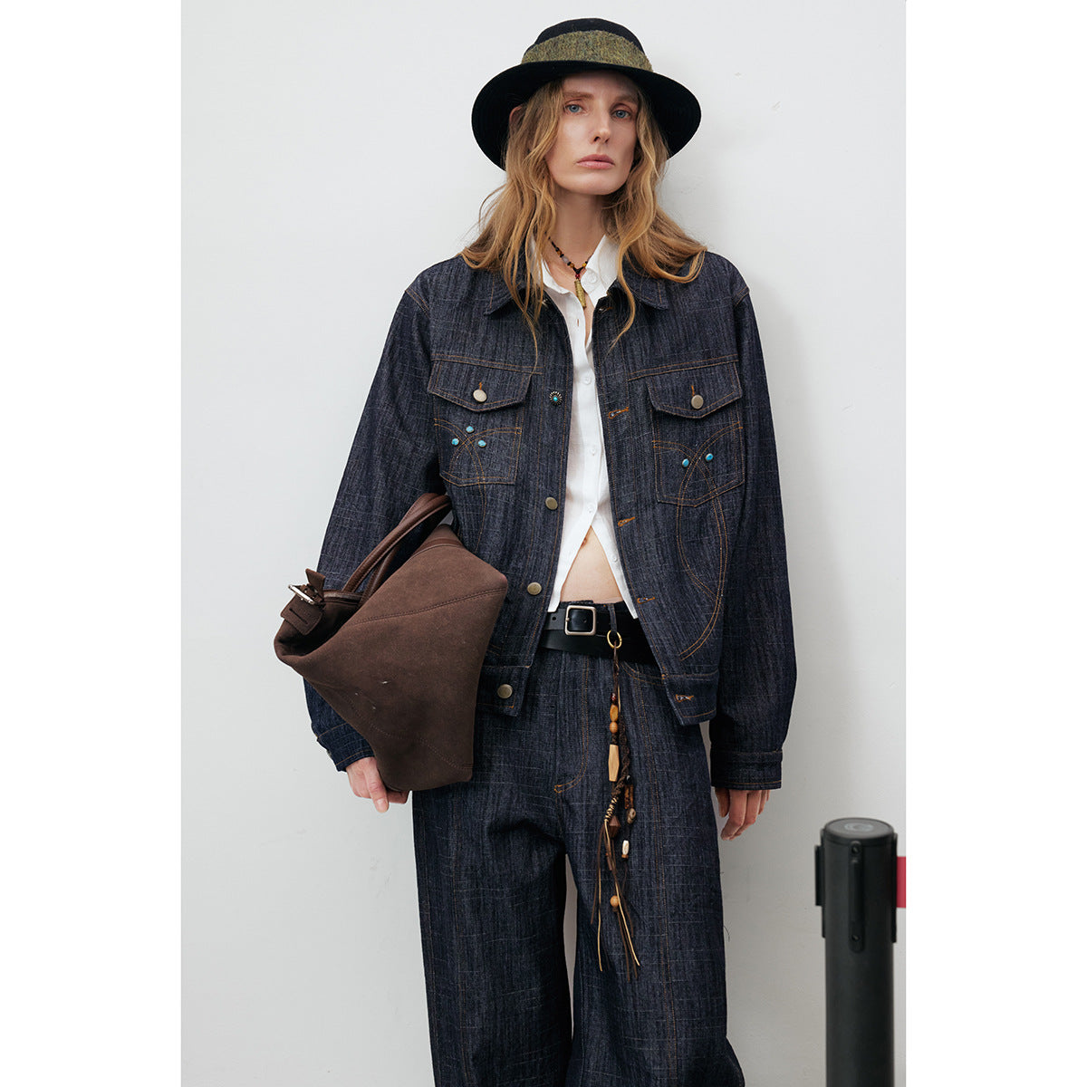 Model wearing an open dark indigo jacket with decorative stitching and matching trousers, holding a brown bag.