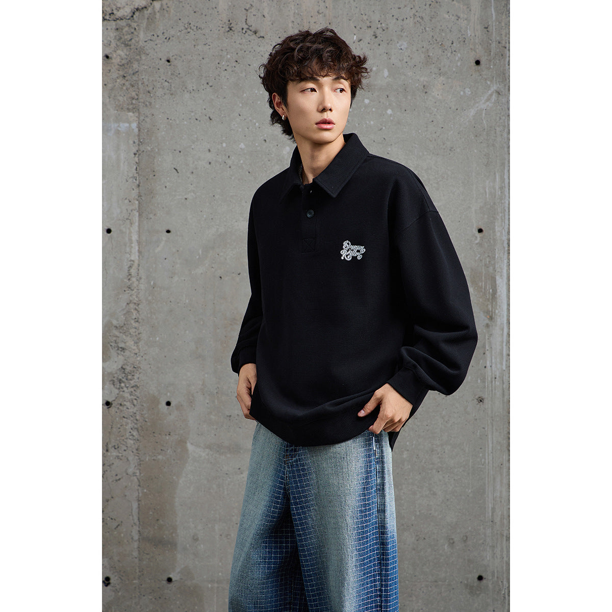 Model wearing a loose black collared pullover with chest embroidery and blue-gray checked trousers.
