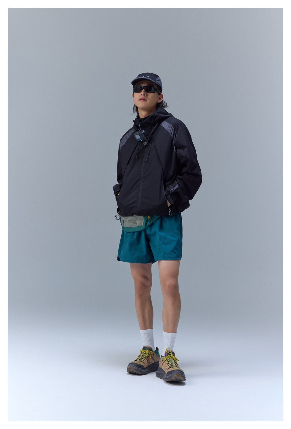 Full-length view of a model wearing a black hooded jacket with gray shoulder panels and teal shorts.