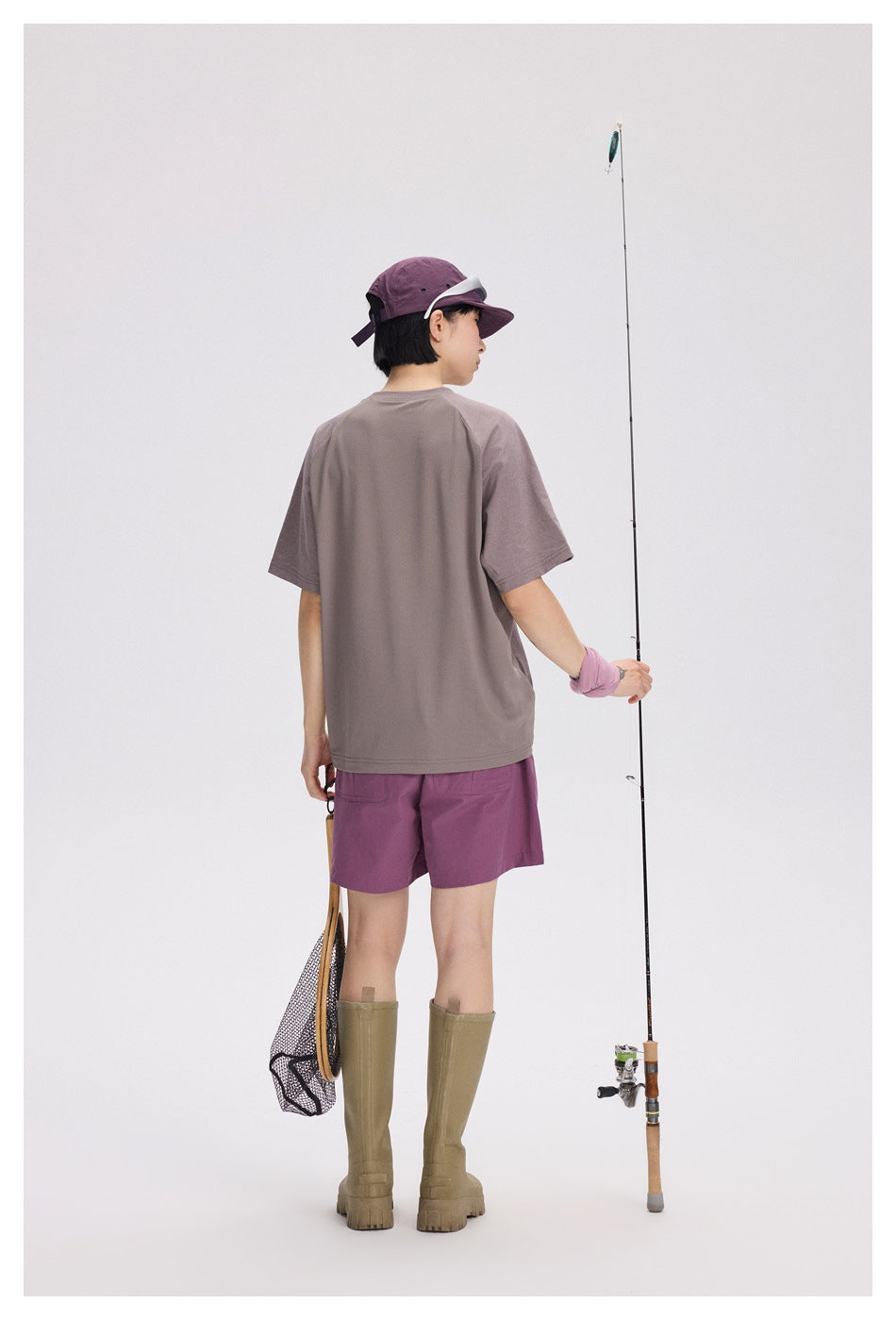 Rear view of a model wearing a mauve T-shirt, purple shorts, purple cap, and olive boots with fishing gear.