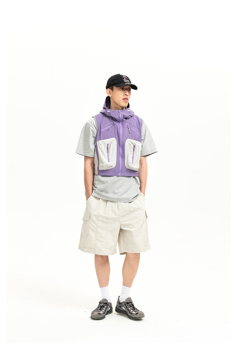 Front view of a model wearing a purple utility vest with white pockets, gray T-shirt, and cream cargo shorts.